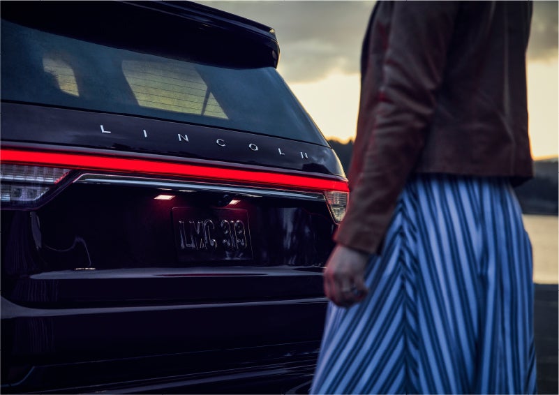 A person is shown near the rear of a 2023 Lincoln Aviator® SUV as the Lincoln Embrace illuminates the rear lights | Midland Lincoln in Midland MI