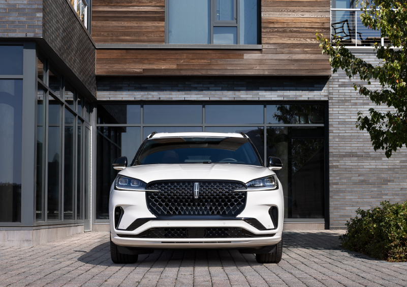 A white 2025 Lincoln Aviator® SUV is shown parked outside a modern building with gray brick and wood accents. | Midland Lincoln in Midland MI