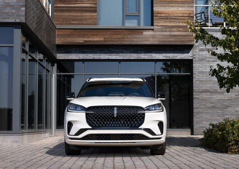 A white 2025 Lincoln Aviator® SUV is shown parked outside a modern building with gray brick and wood accents. | Midland Lincoln in Midland MI