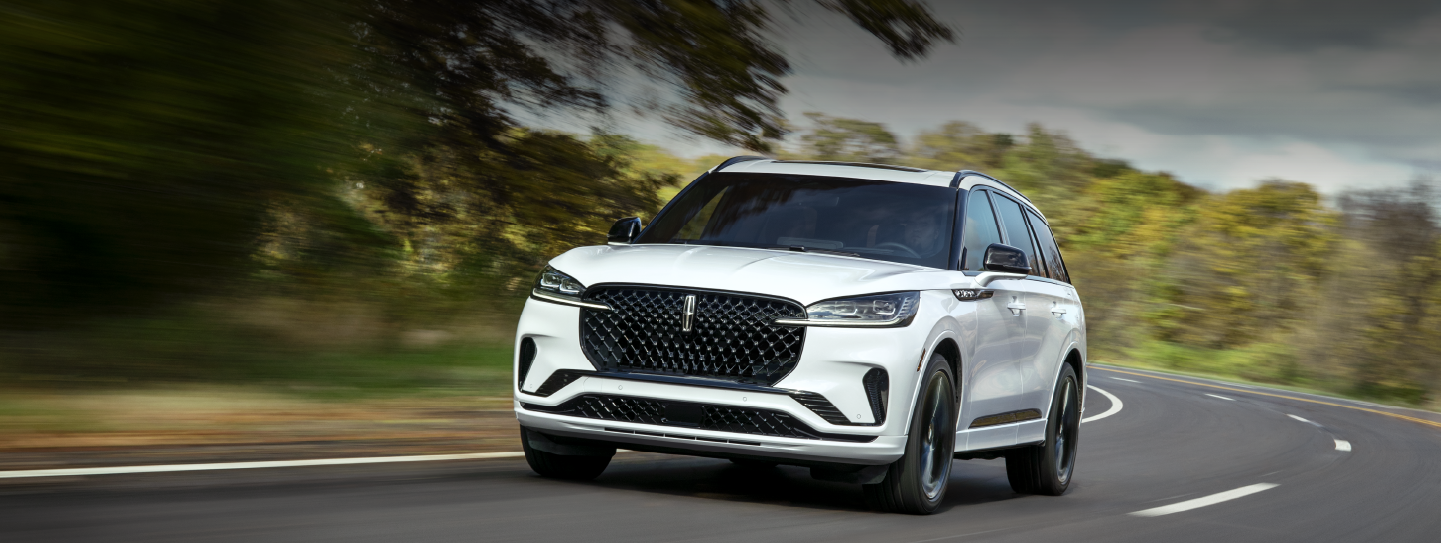 A white 2025 Lincoln Aviator® SUV is being driven down a road lined with trees. | Midland Lincoln in Midland MI