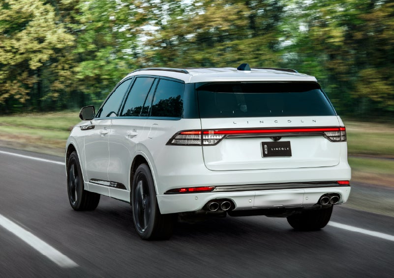 A white 2026 Lincoln Aviator® SUV is being driven away down a road lined with colorful trees. | Midland Lincoln in Midland MI
